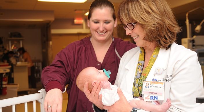 One nurse holds baby in NICU while a second nurse looks on